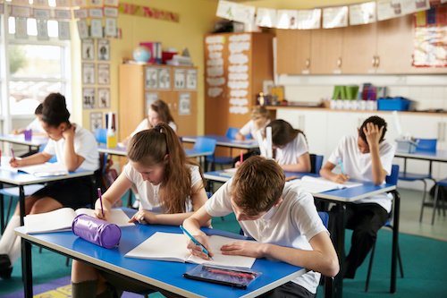 Students working at their desks in a bright classroom, writing in their notebooks.