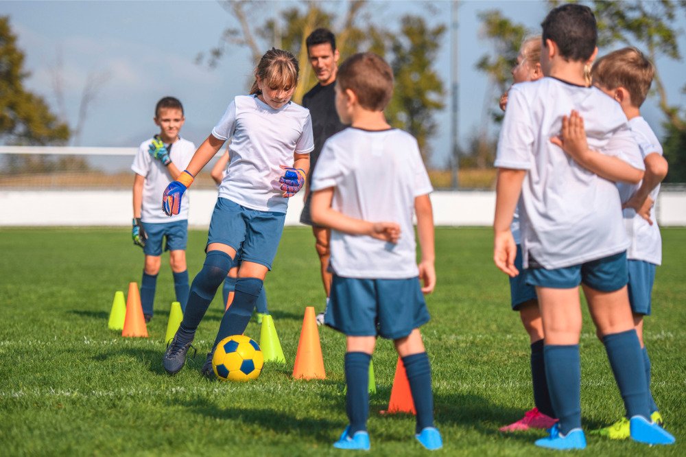 Children in football kit dribbling a ball between training cones on a grass pitch while their coach looks on.